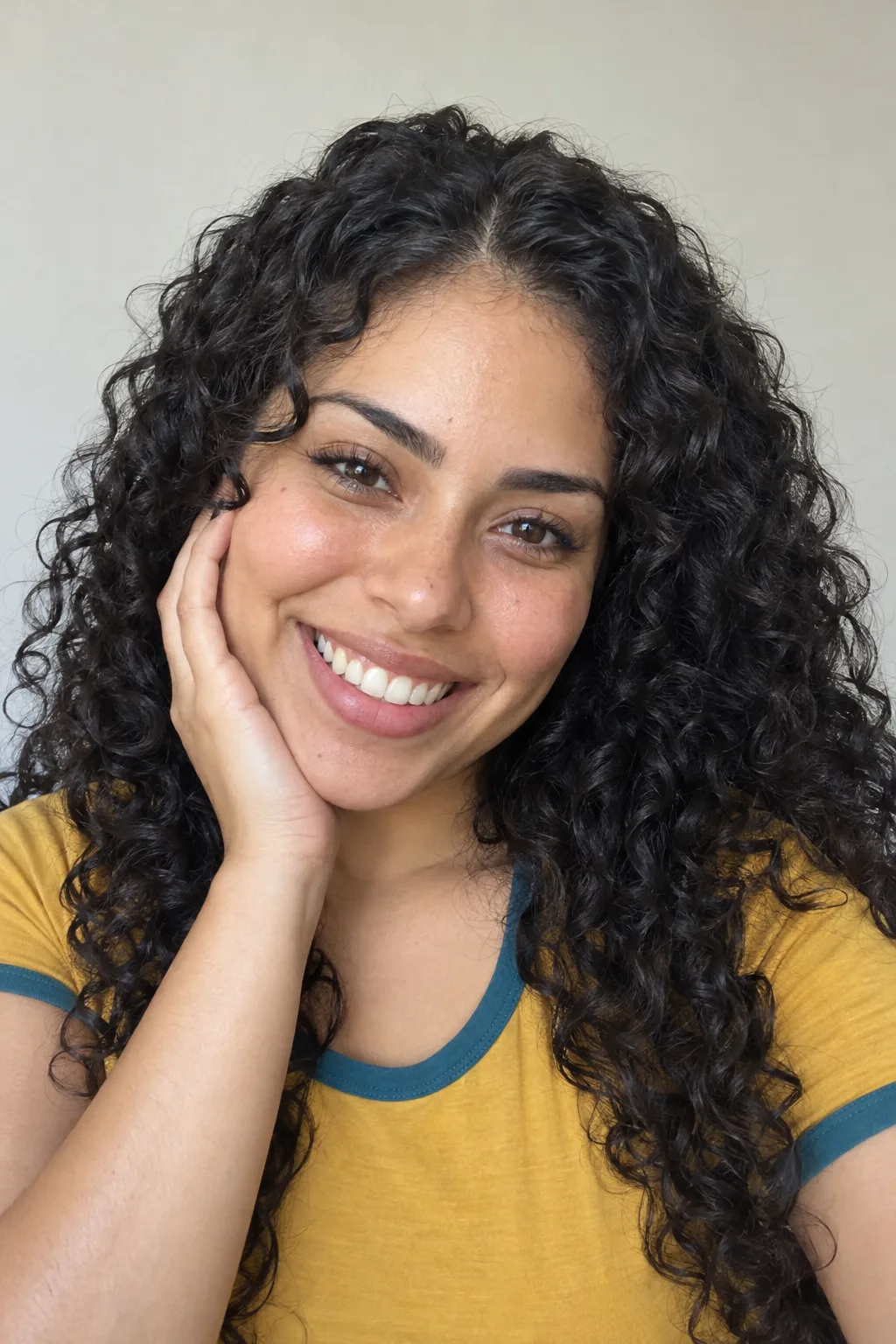 Adult with black ringlets, smiling brightly with one hand beside her cheek.