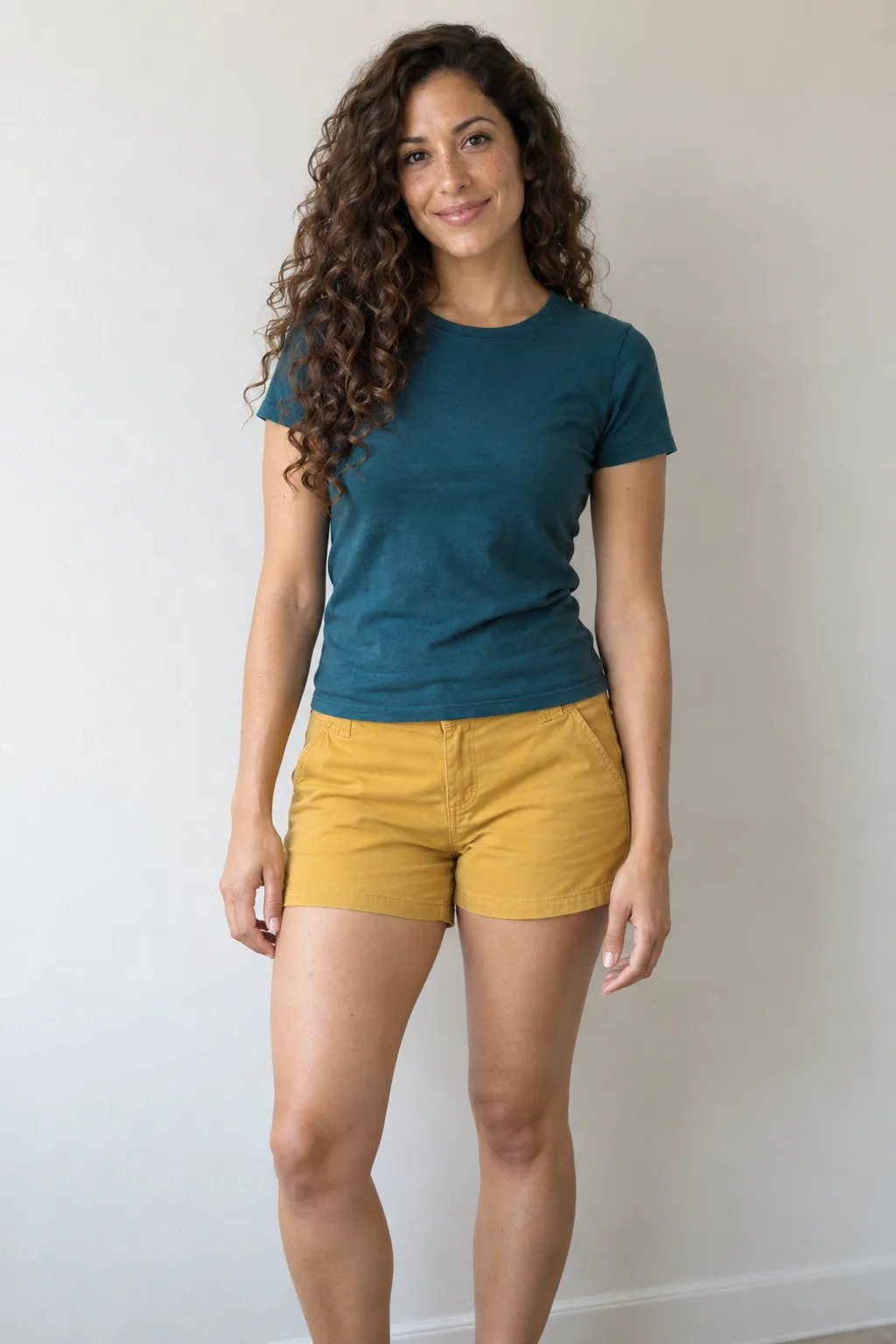 Adult with long chestnut curls standing in a teal T-shirt and mustard shorts.